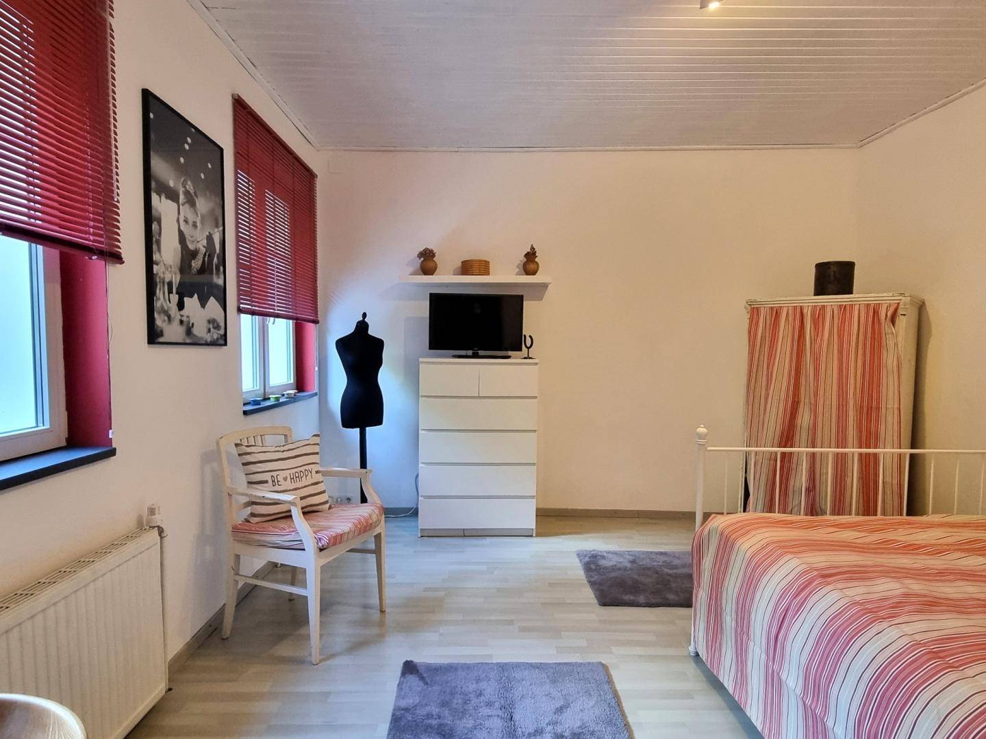 Schlafzimmer mit hellem Holzboden, weißem Bett und roten Jalousien, einfach und funktional eingerichtet.