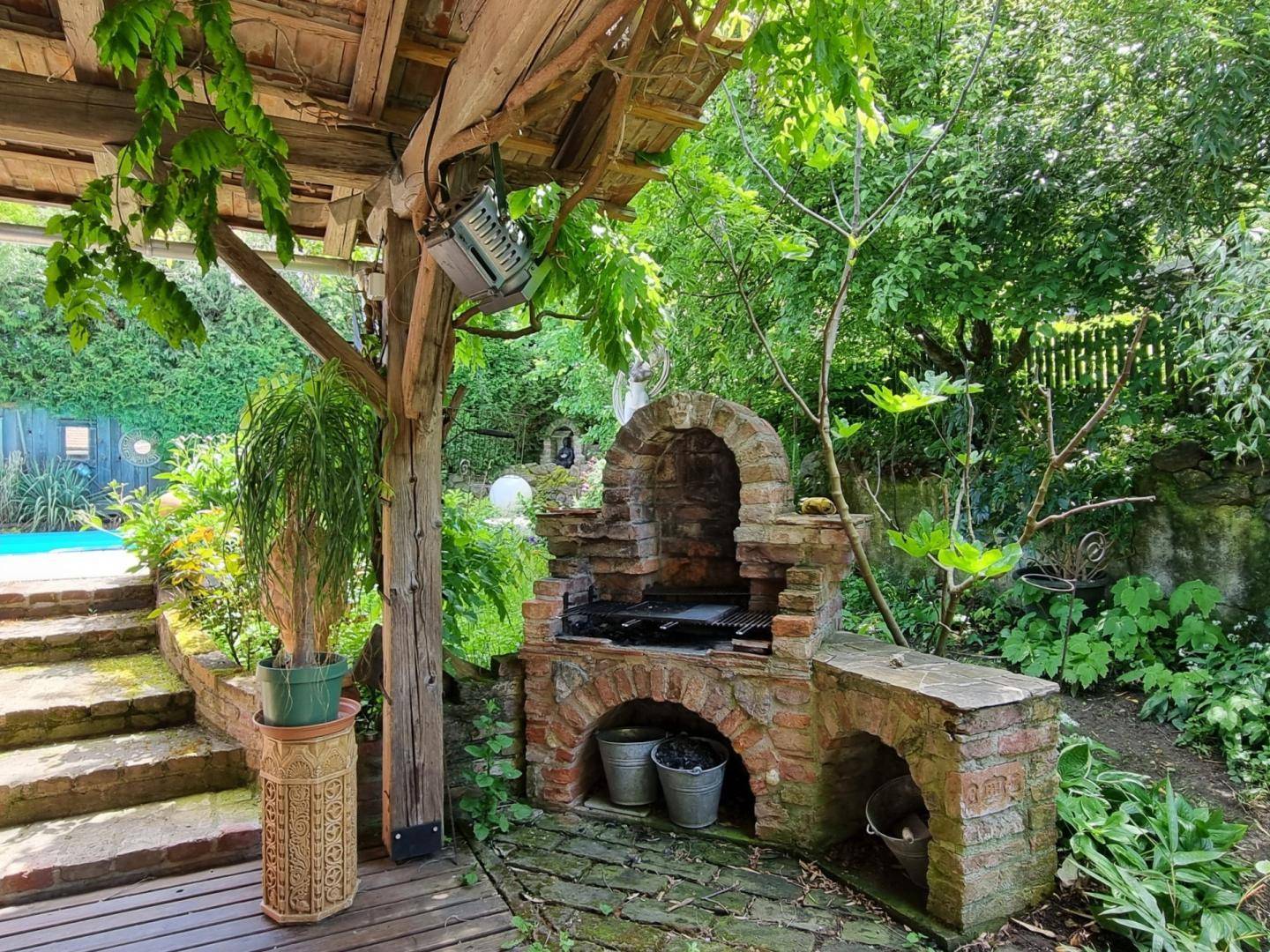Gemauerter Grillplatz im Garten unter einer Holzpergola, umgeben von üppiger Vegetation.