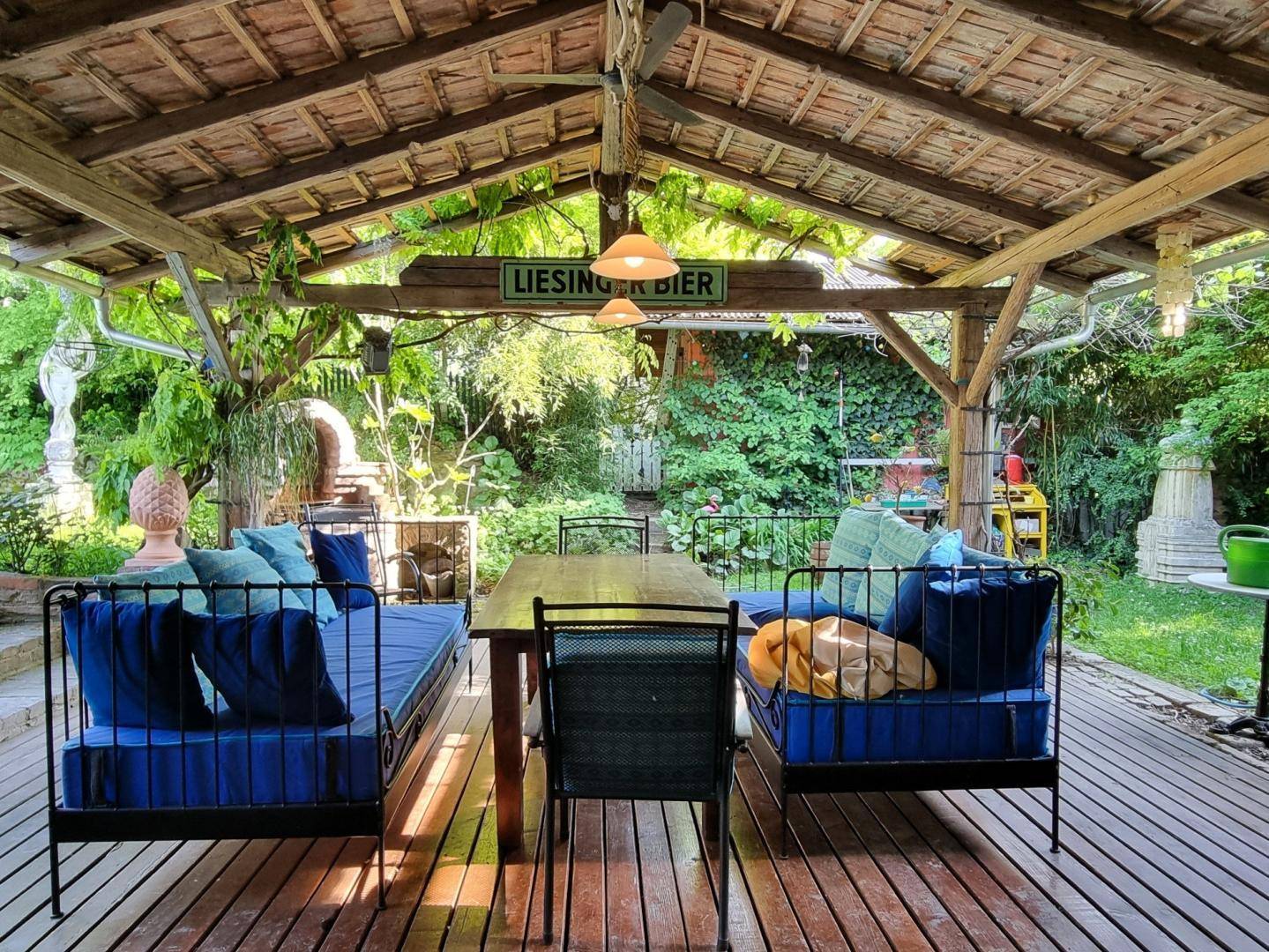 Überdachte Holzterrasse mit bequemen blauen Sitzmöbeln und Esstisch, umgeben von üppigem Grün.