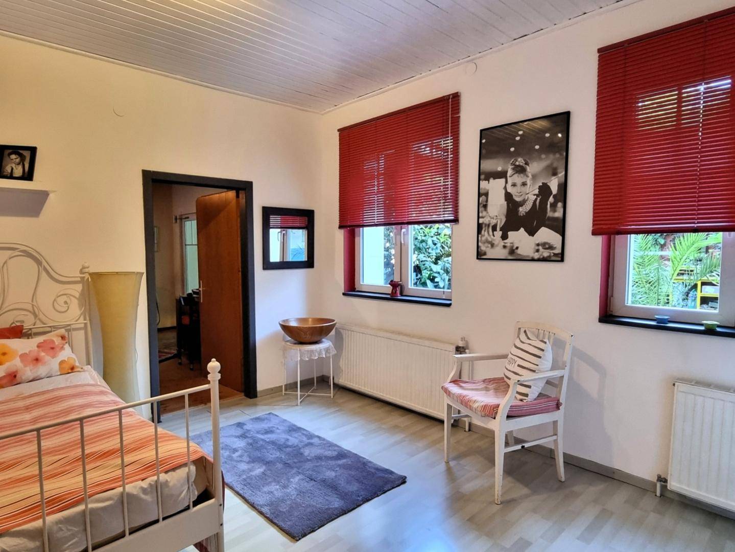 Schlafzimmer mit Holzboden, weißem Bett und roten Jalousien, mit Zugang zu einem weiteren Raum.