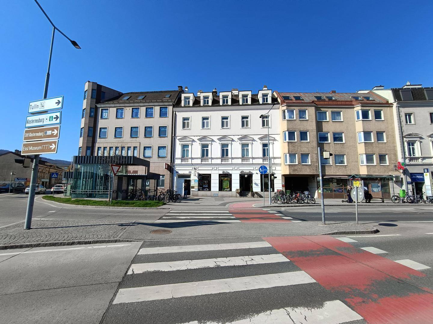Stadtansicht mit einer Reihe von Gebäuden, einem Zebrastreifen und Verkehrsschildern unter blauem Himmel.