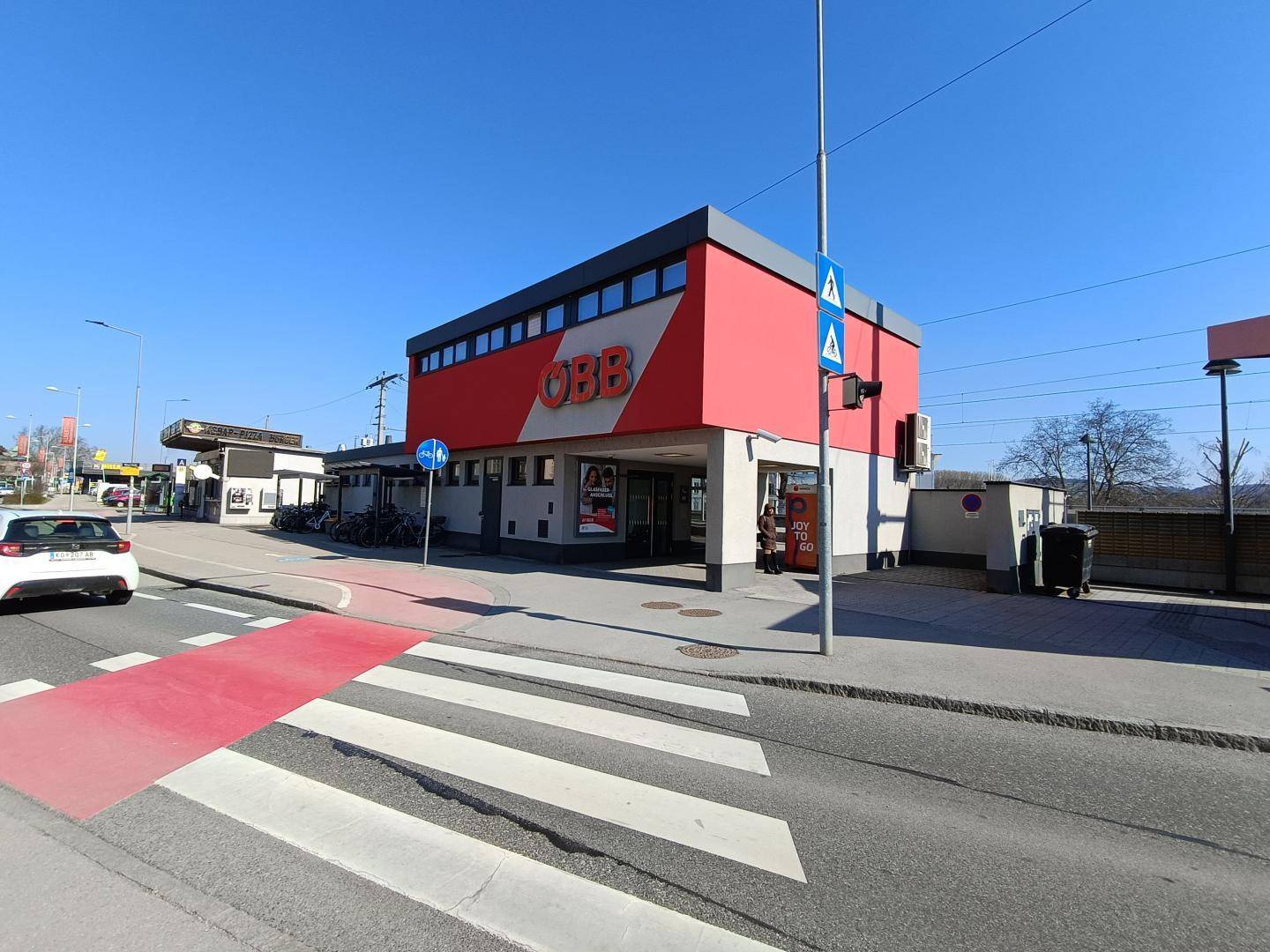 Straßenansicht mit einem ÖBB-Gebäude, einem Zebrastreifen und einer Tankstelle im Hintergrund, städtische Umgebung.