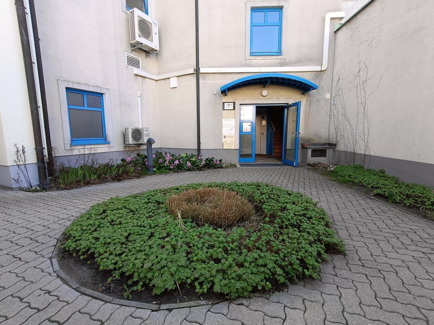 Eingangsbereich eines Gebäudes mit gepflastertem Hof, Pflanzen und blauen Fensterrahmen, einladend gestaltet.