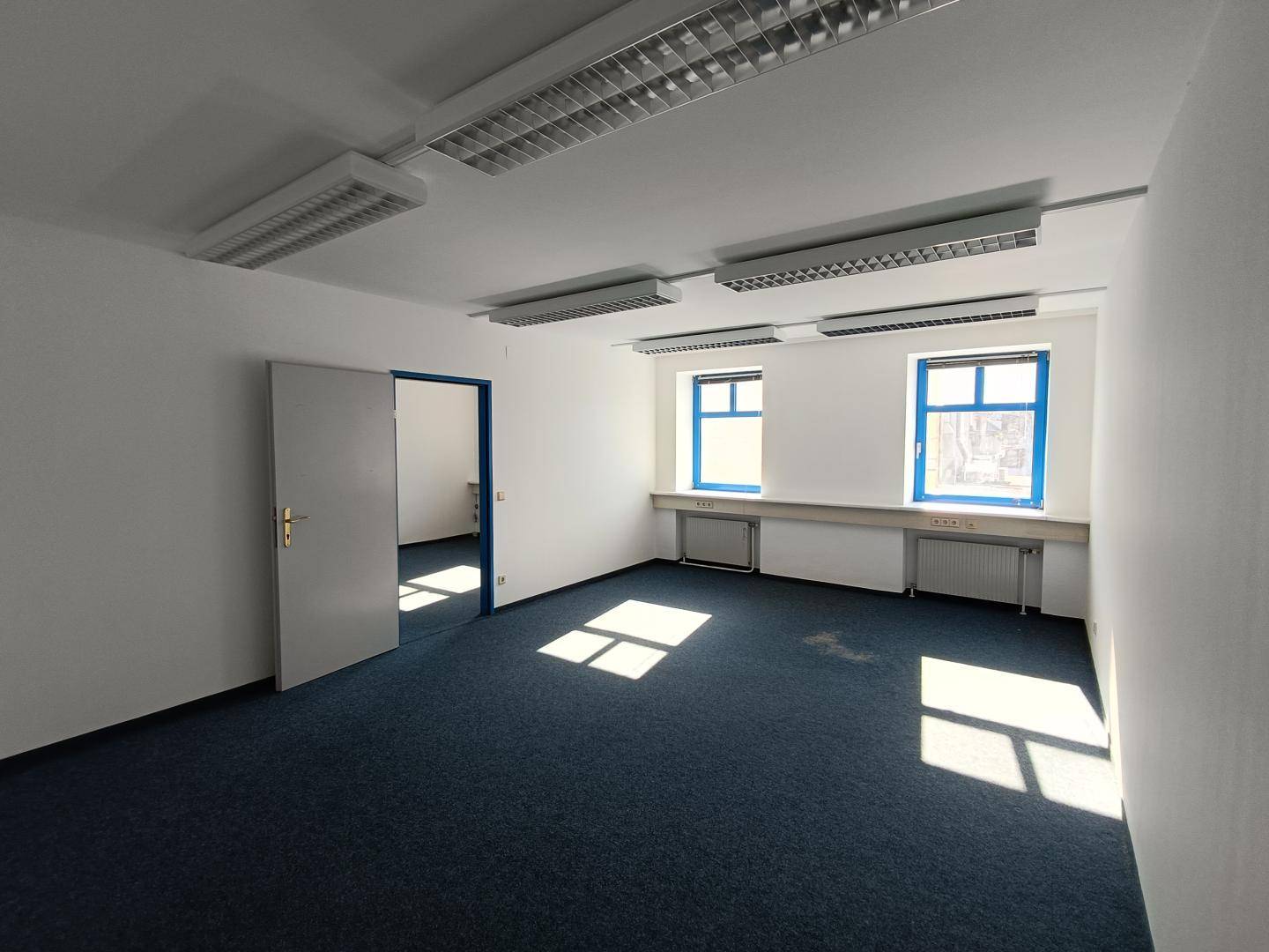 Geräumiges, leeres Büro mit blauem Teppichboden und Zugang zu weiteren Räumen, hell und sauber.