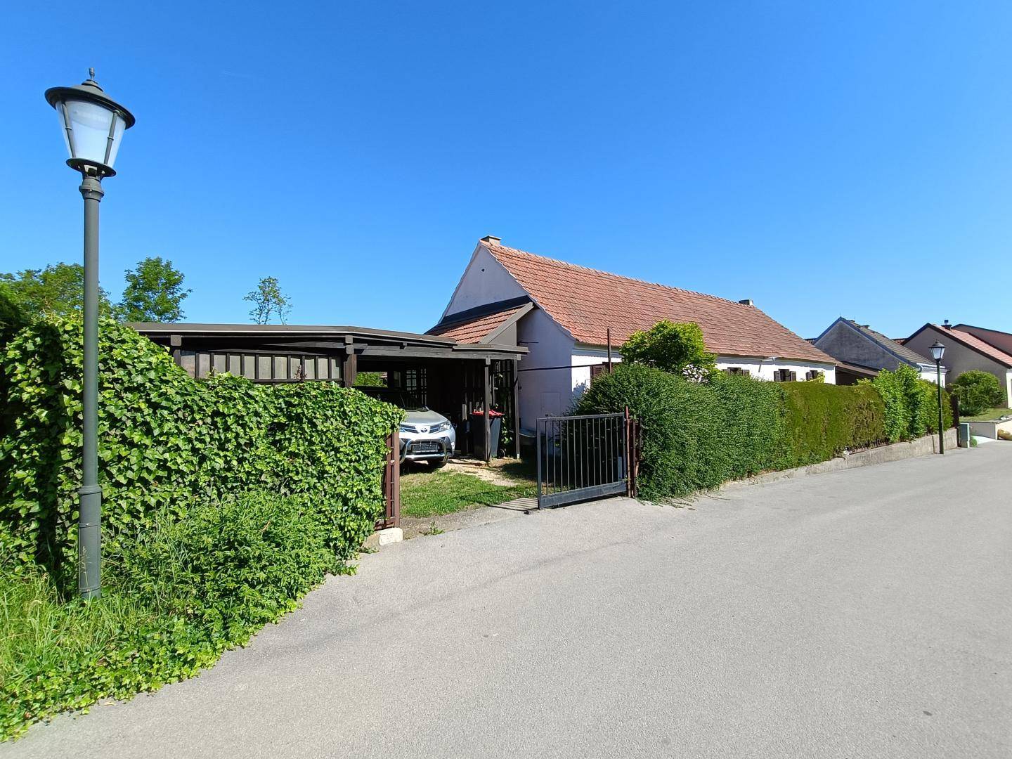 Ansicht des Landhauses von der Straße mit einem begrünten Carport und einem Gartentor.