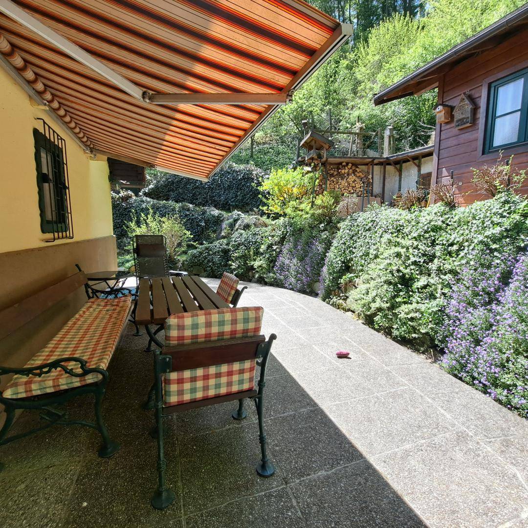 Gepflasterte Terrasse mit Markise, Gartenmöbeln und üppiger Bepflanzung im Hintergrund.