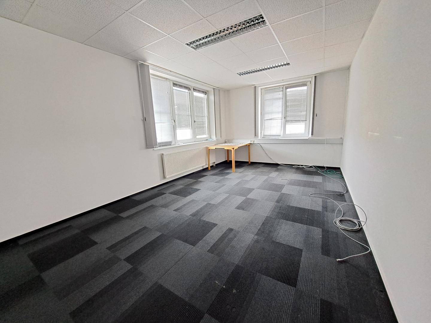 Leeres Büro mit Teppichboden, weißen Wänden und einem kleinen Holztisch unter den Fenstern.