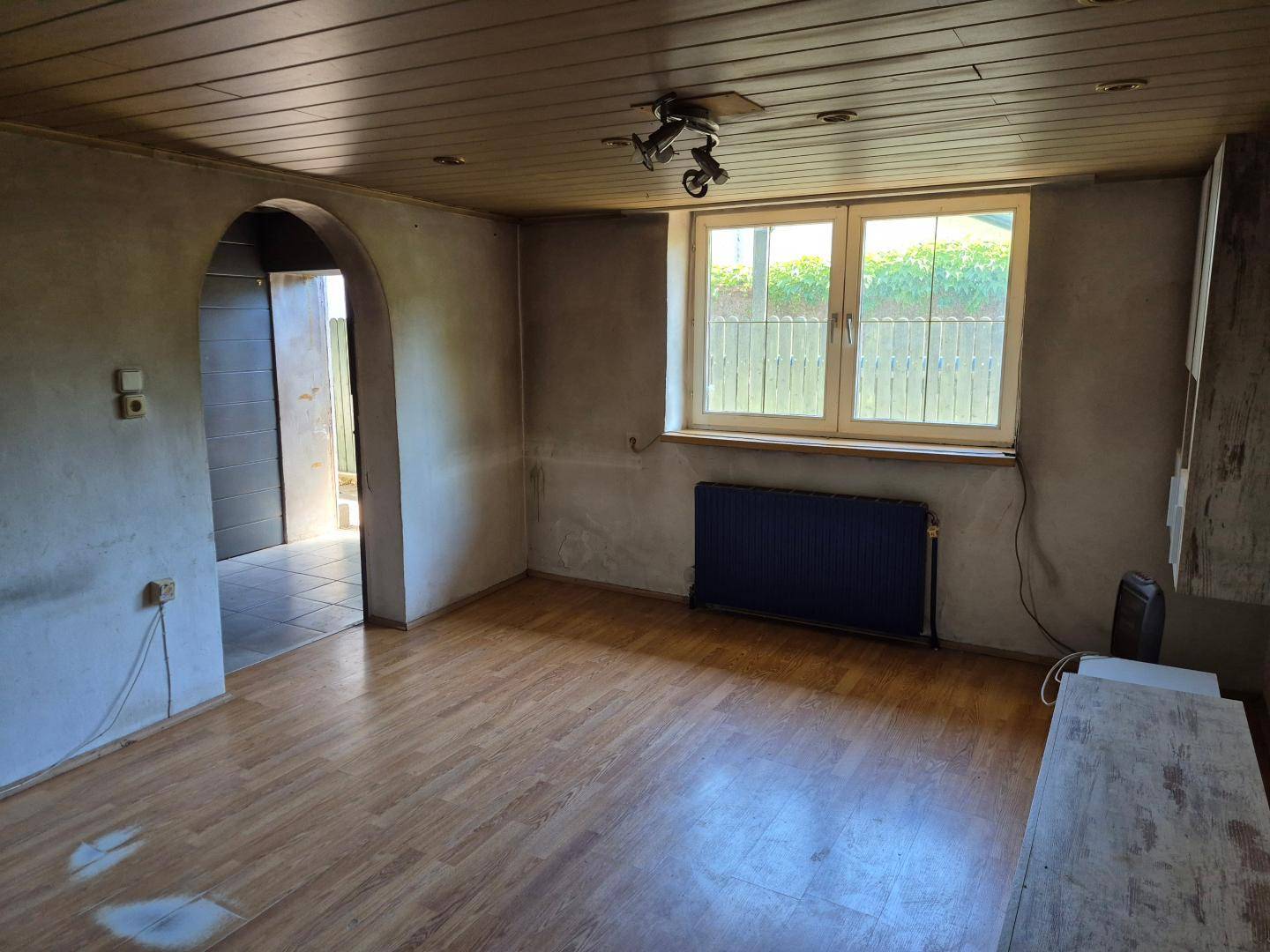 Renovierungsbedürftiger Wohnraum mit Fenster, Heizkörper und Holzbalkendecke.