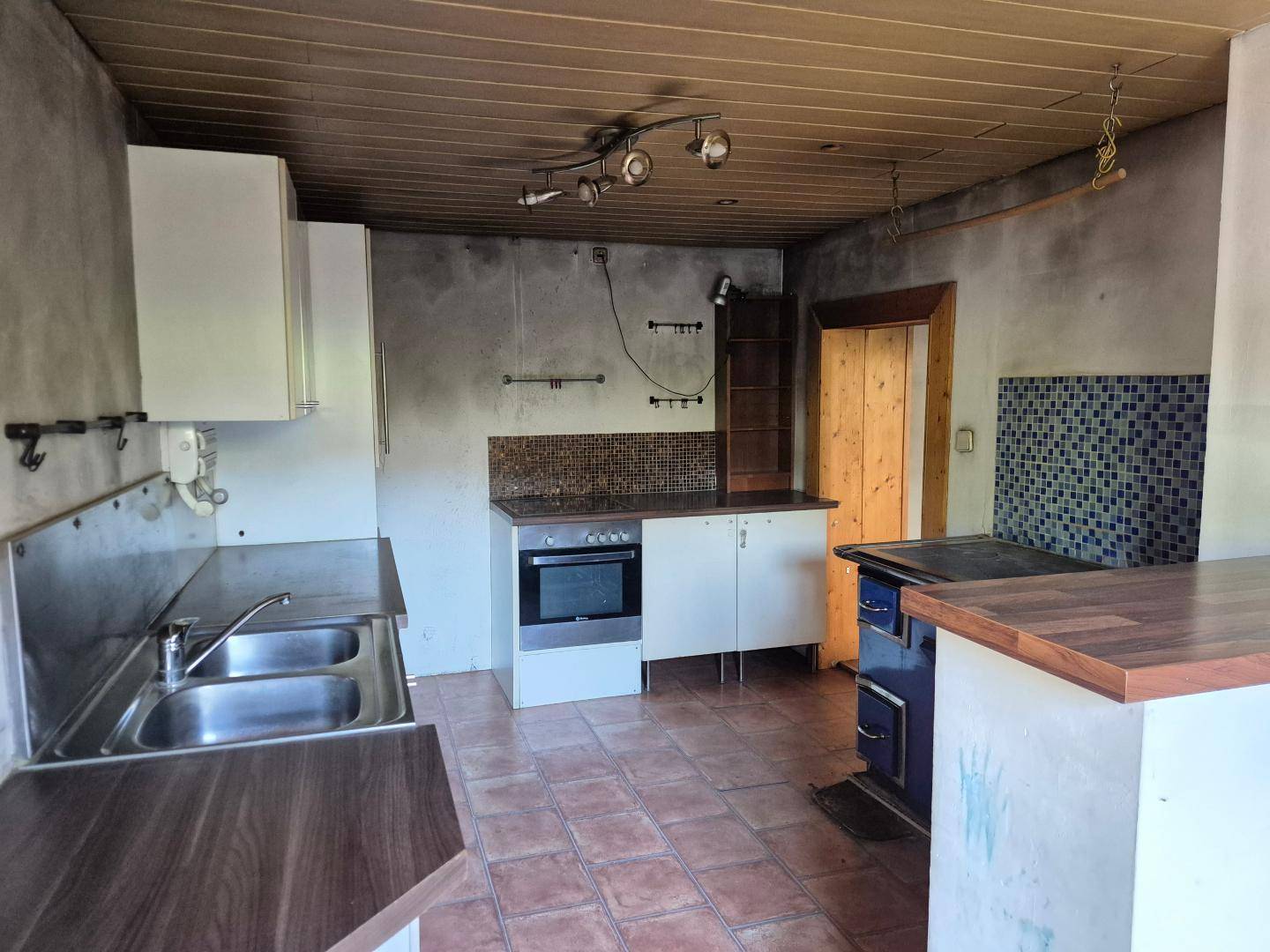 Rustikale Küche mit blauer Ofenbank, Doppelspüle und Holzdecke, renovierungsbedürftig.