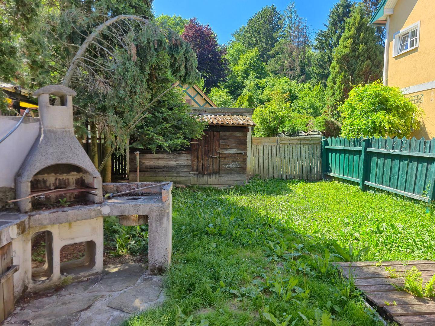 Gartenbereich mit gemauertem Grill und Holzschuppen, umgeben von üppigem Grün.