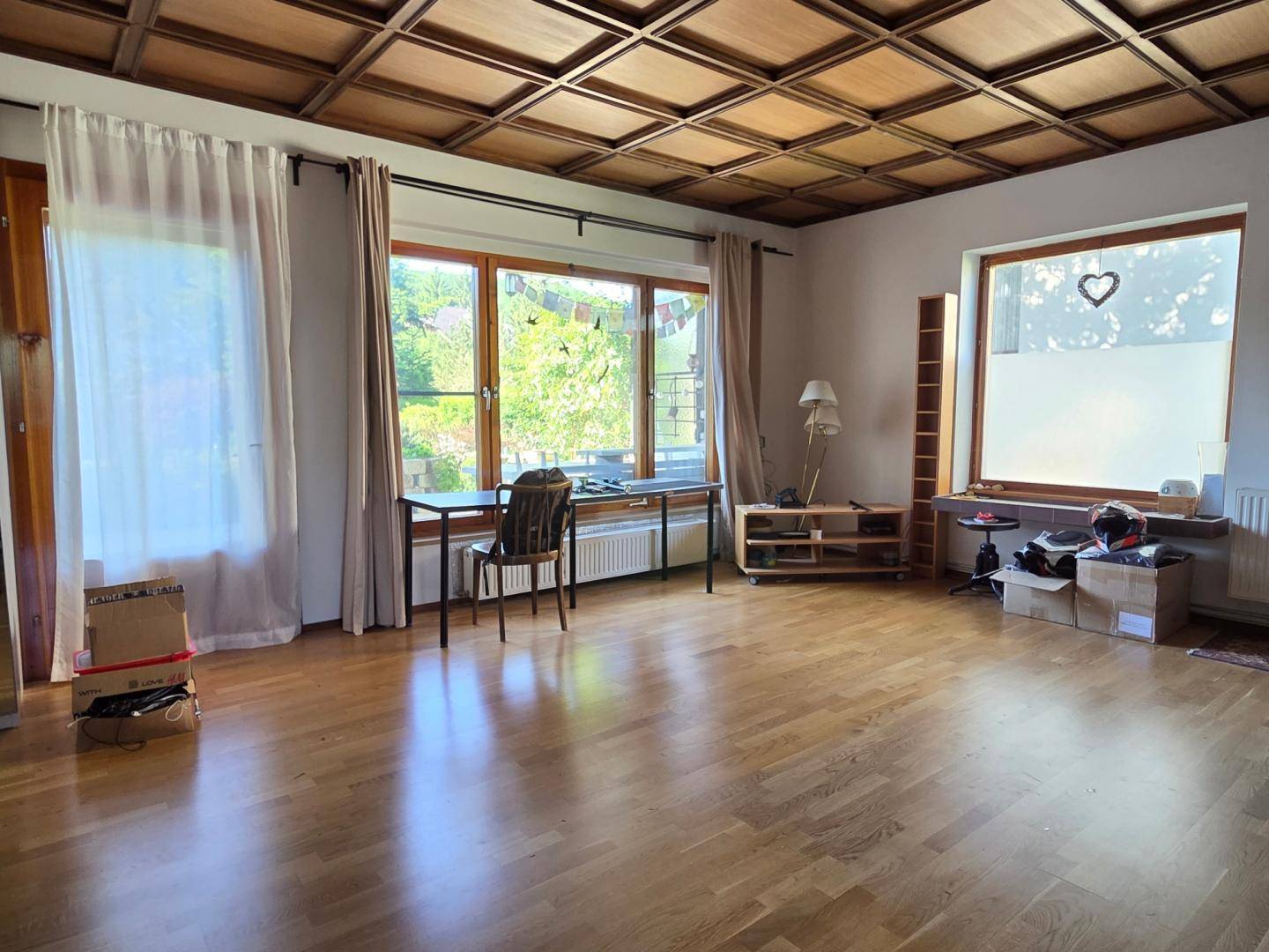 Helles Wohnzimmer mit Holzboden, Kassettendecke und großen Fenstern mit Blick ins Grüne.