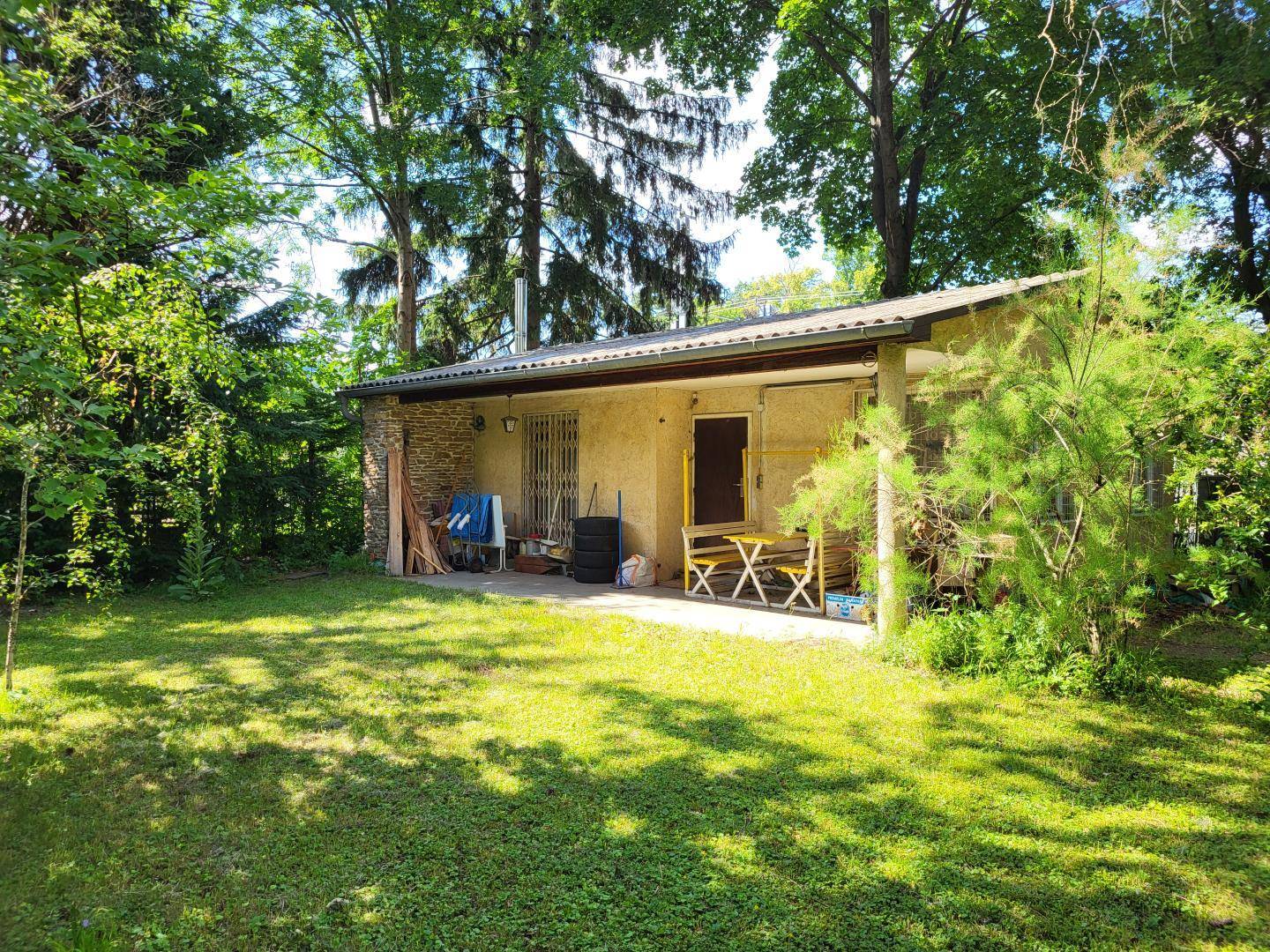 Kleines Haus mit überdachter Terrasse und Gartenmöbeln, umgeben von viel Grün.