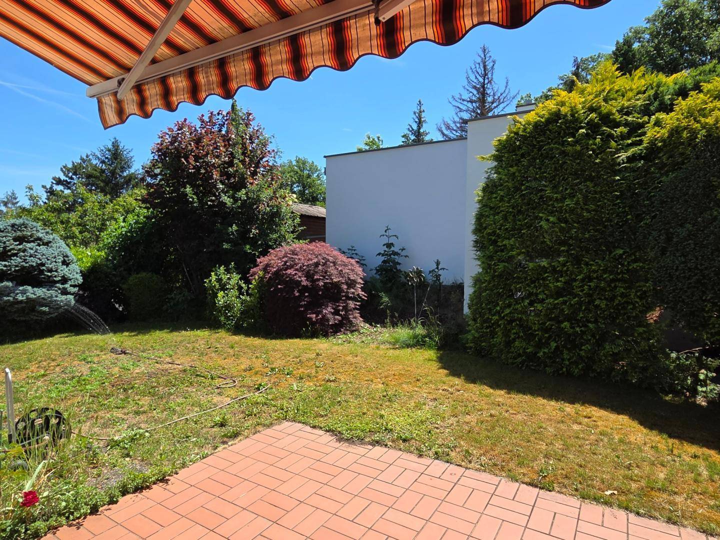 Sonniger Gartenbereich mit Rasenfläche und einer kleinen gepflasterten Terrasse unter einer Markise.