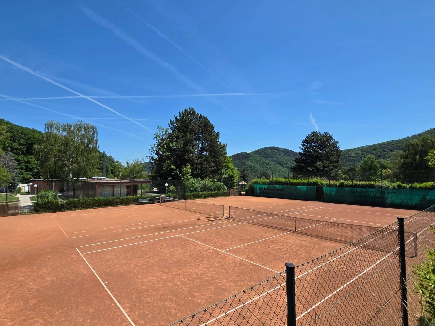 Weitläufige Tennisplätze mit rotem Sand, eingebettet in eine grüne Hügellandschaft unter blauem Himmel.
