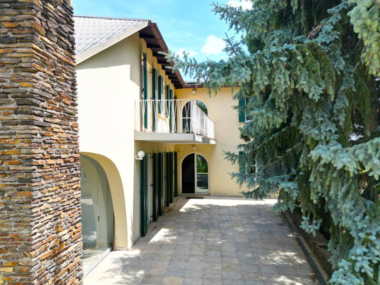 Außenansicht des Hauses mit gepflastertem Weg, Steinkamin und einem Balkon im Obergeschoss.
