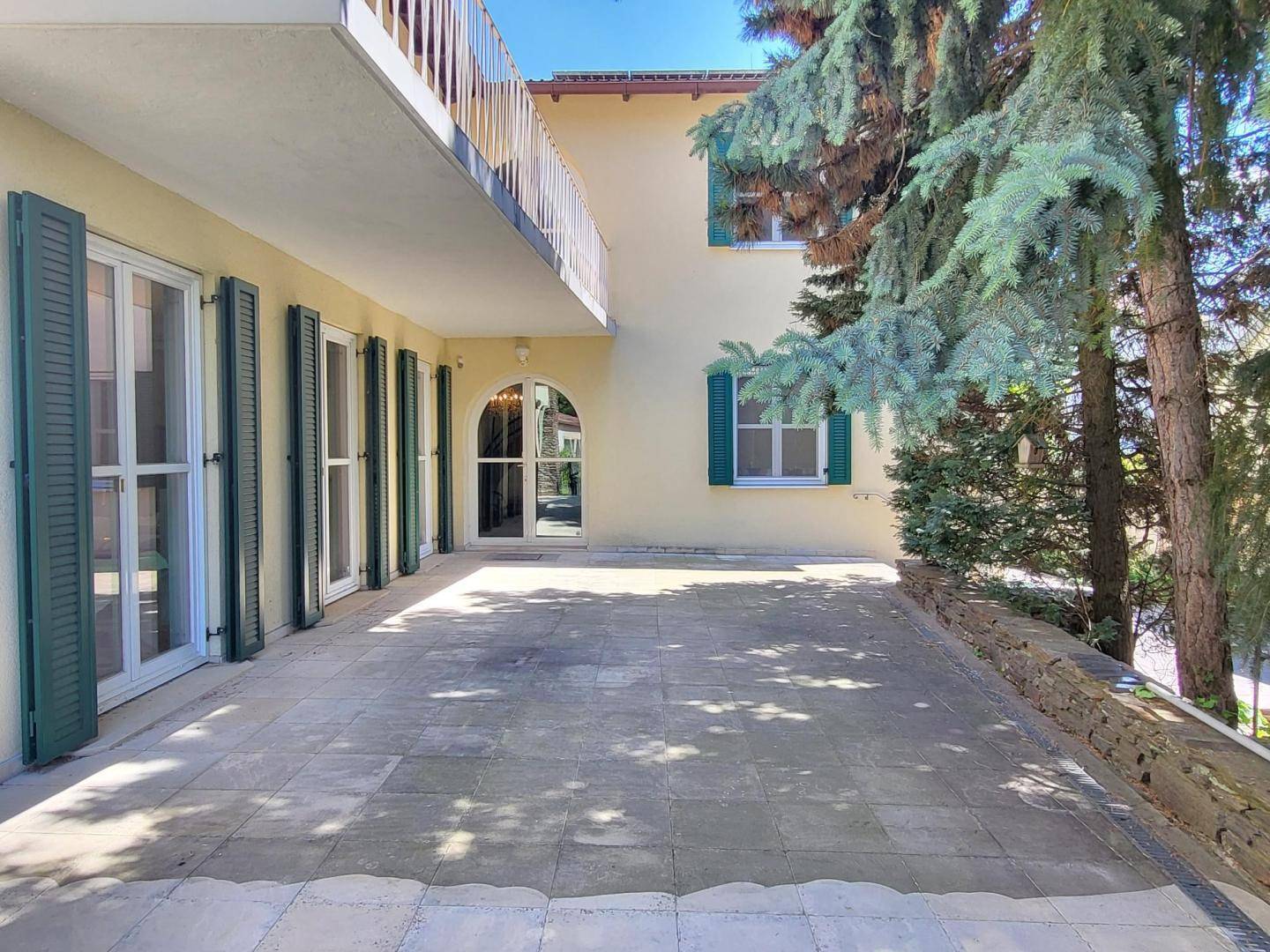 Weitläufige gepflasterte Terrasse vor dem Haus mit grünen Fensterläden und einem Balkon darüber.