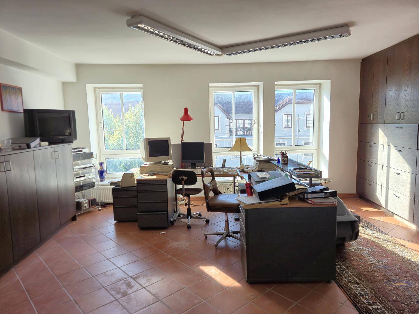Büro mit älterer Ausstattung, zahlreichen Schränken und einem Teppich auf dem Fliesenboden.
