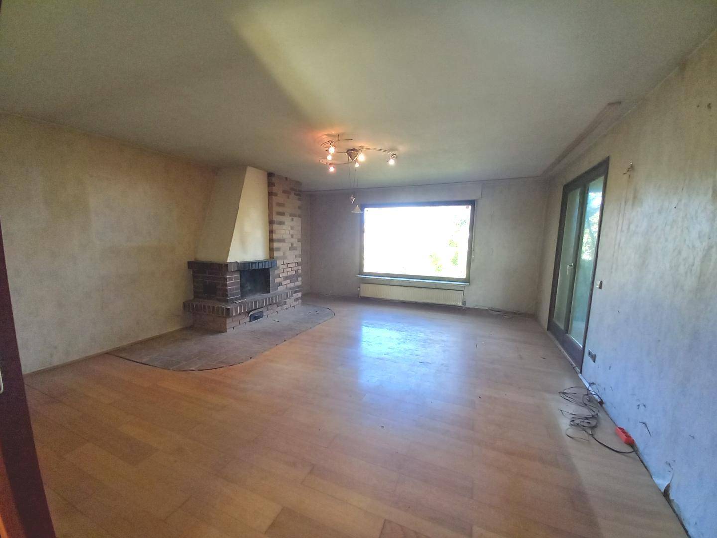 Geräumiges Wohnzimmer mit Holzboden, gemauertem Kamin und großem Fenster.