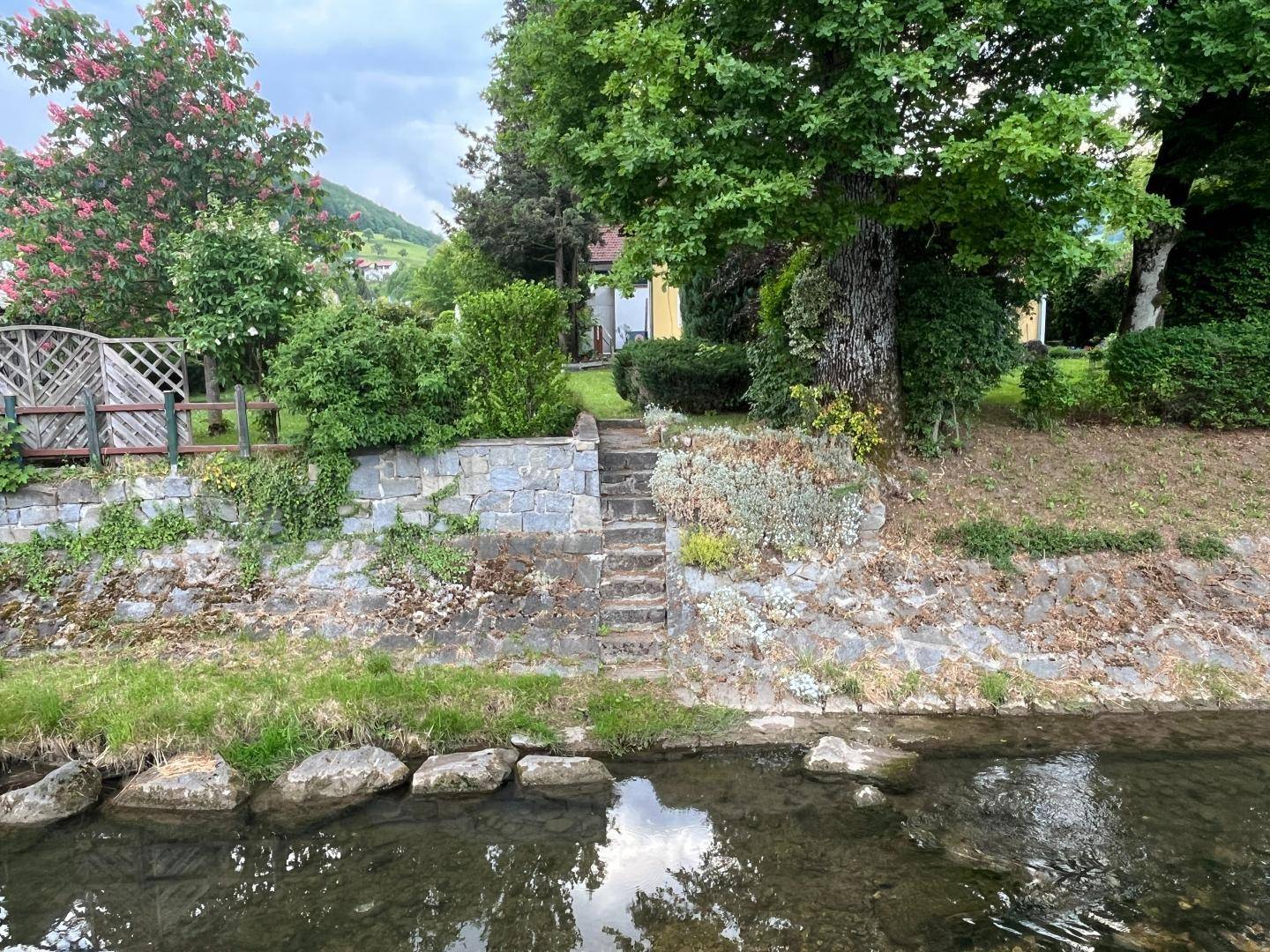 Blick auf das Grundstück von einem Bach aus, mit Steintreppe und viel Grün.
