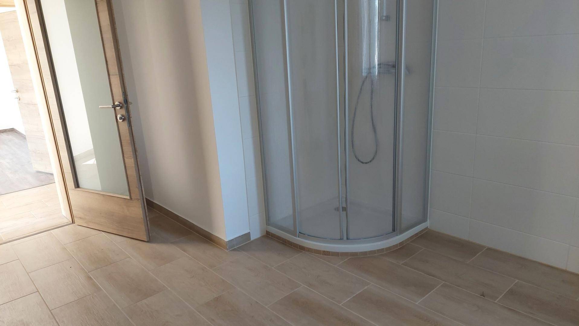 Modernes Badezimmer mit Eckdusche, hellen Fliesen und einer Holztür zu einem weiteren Raum.