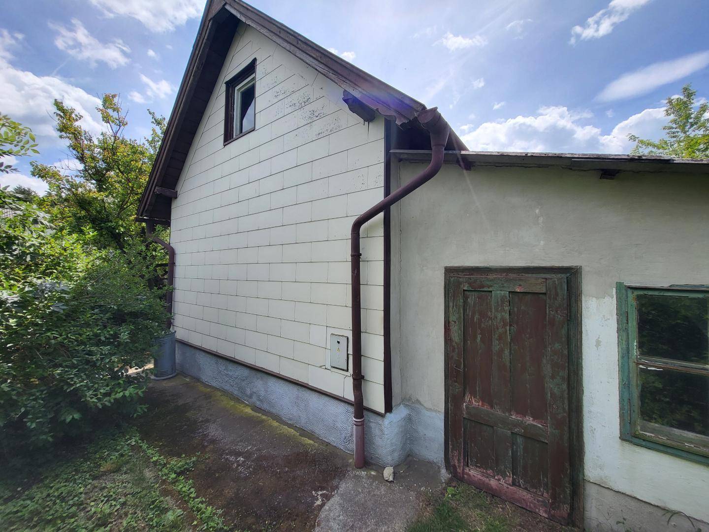 Seitenansicht des Hauses mit verwitterter Holzverkleidung, einem kleinen Fenster und einer alten Holztür.