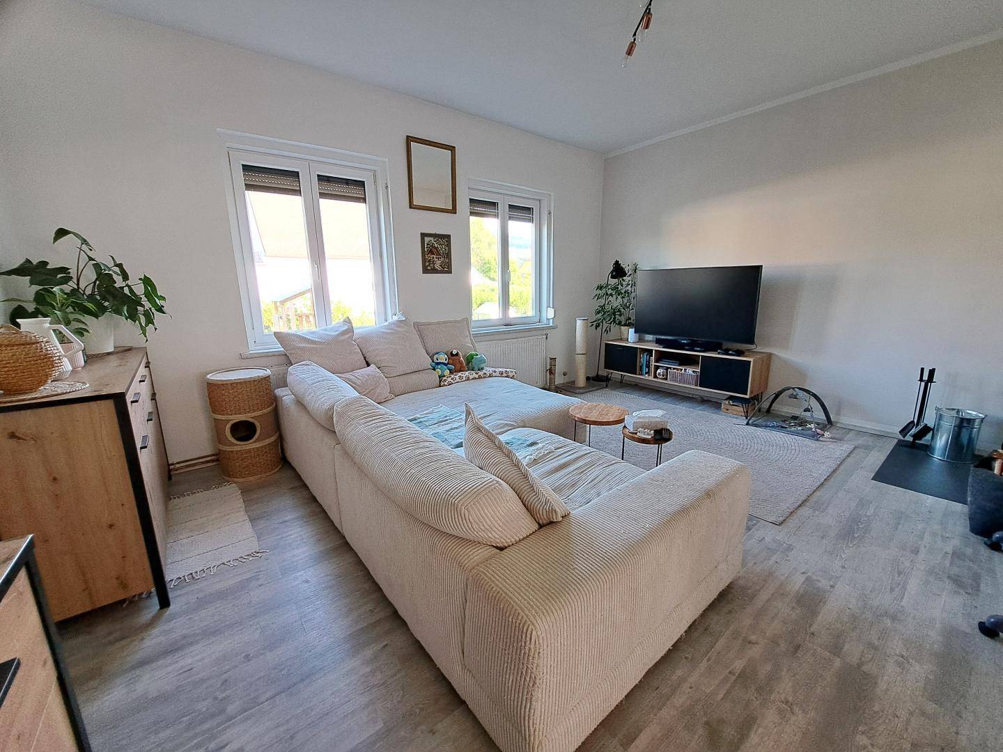 Wohnzimmer mit beigem Sofa, Fernseher und Blick ins Grüne durch die Fenster.