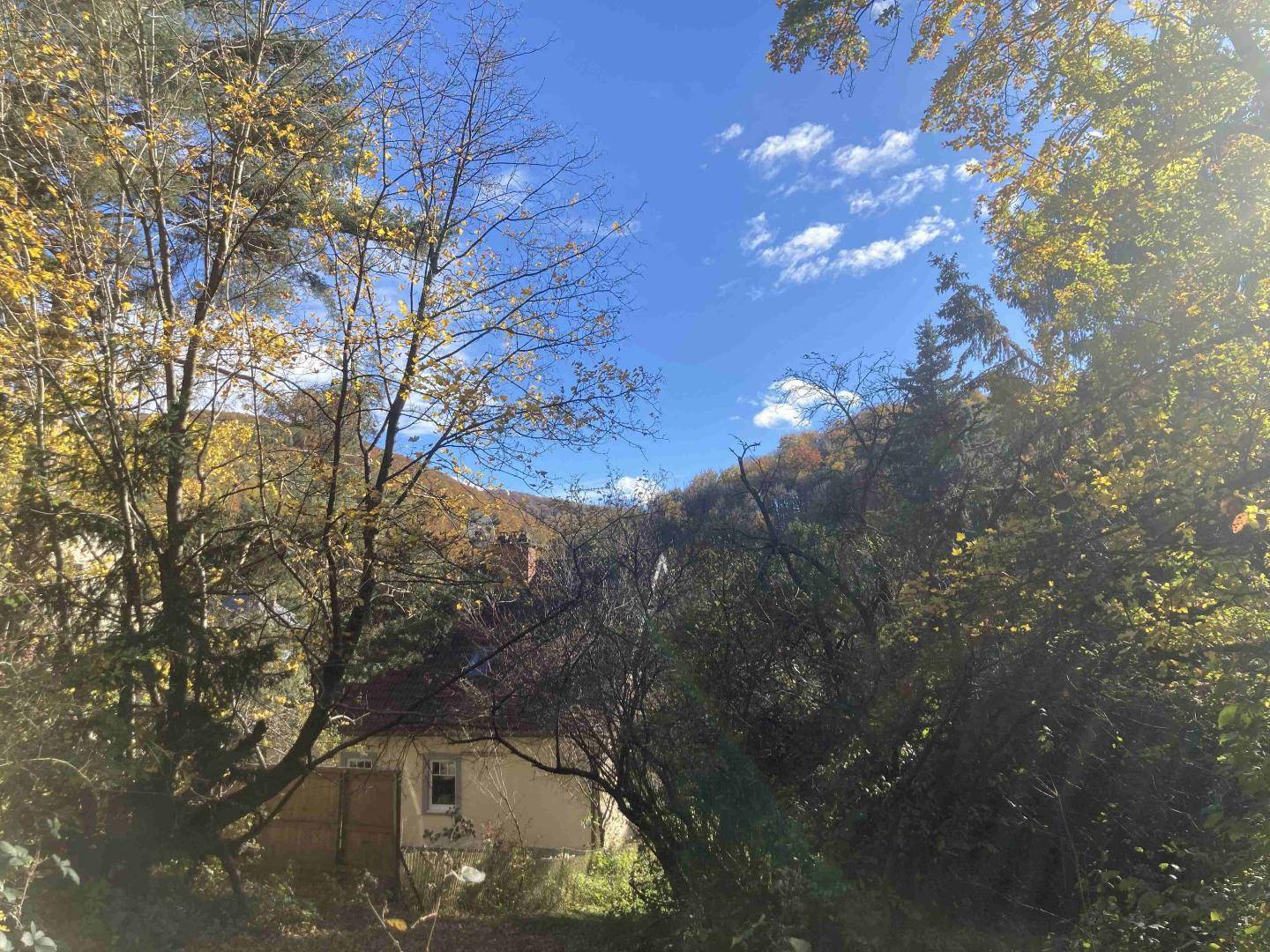 Blick auf ein kleines, älteres Haus umgeben von Bäumen und Hügeln unter einem klaren Herbsthimmel.