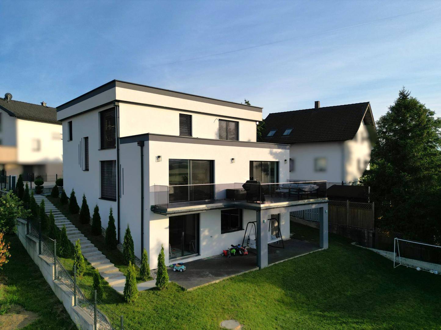Modernes Wohnhaus mit weißer Fassade, Flachdach und Terrassenbereich, umgeben von einem grünen Garten.