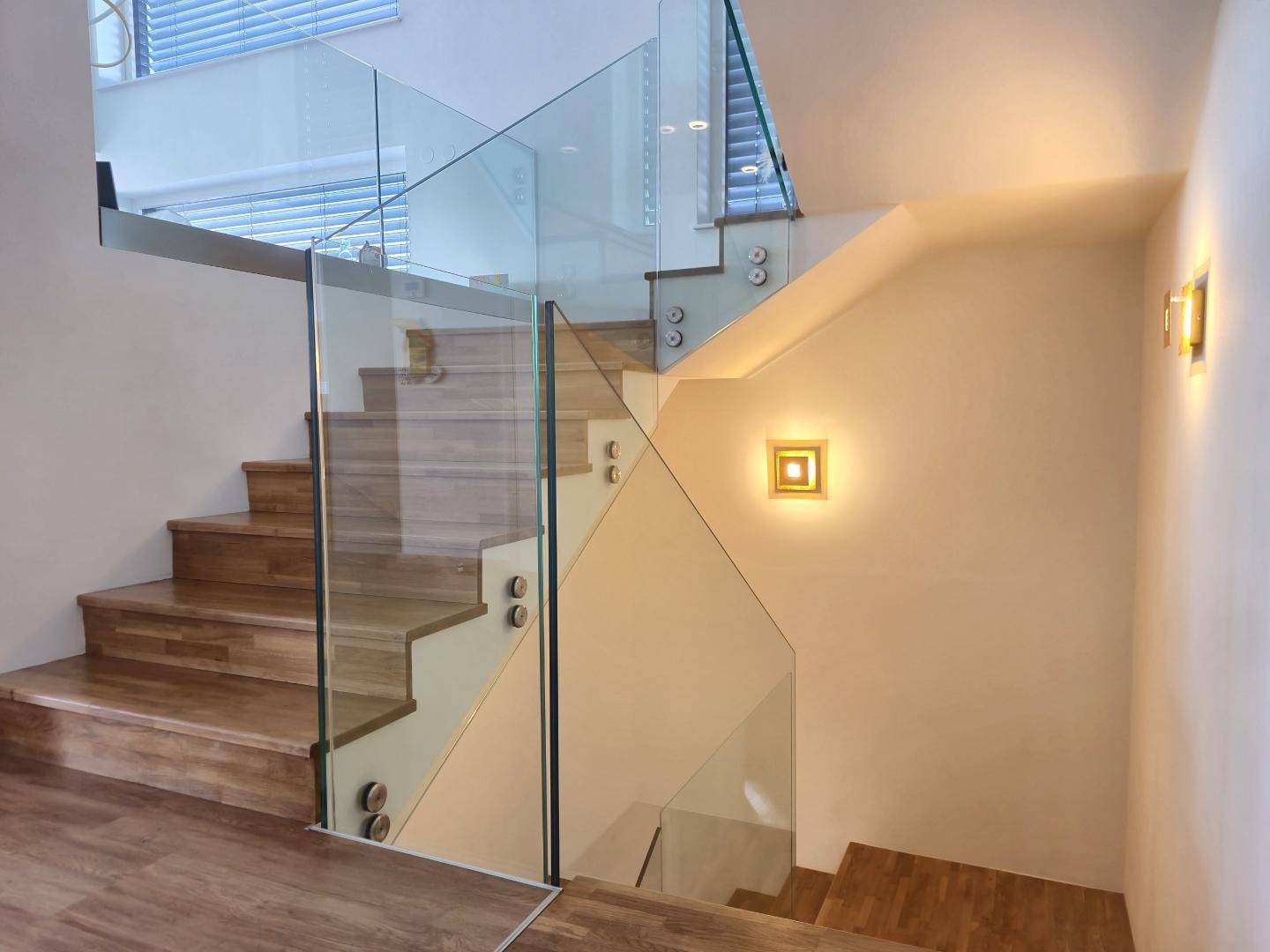 Moderne Holztreppe mit elegantem Glasgeländer und stilvoller Wandbeleuchtung, die den Aufgang erhellt.