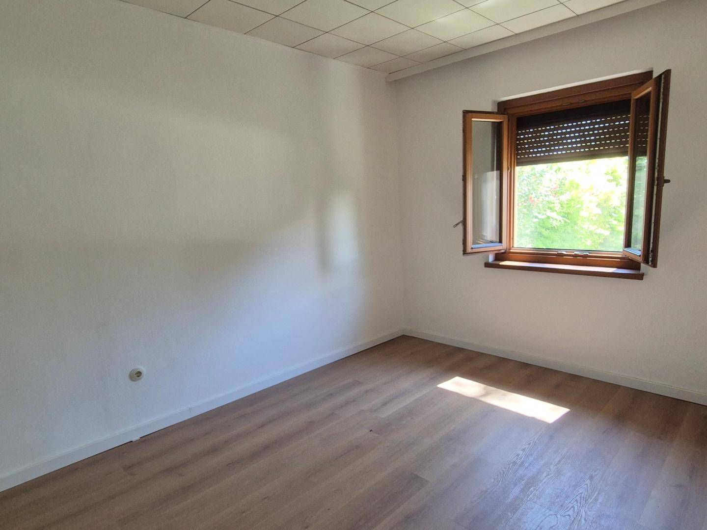 Leeres Zimmer mit Laminatboden, weißen Wänden und einem Fenster mit Rollladen.