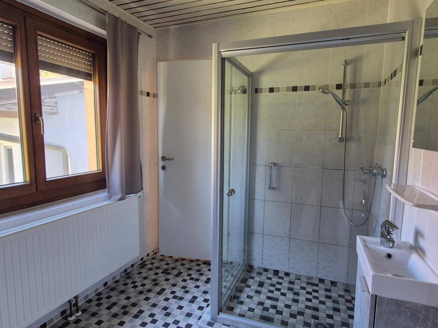 Badezimmer mit Duschkabine, kleinem Waschbecken und auffälligen schwarz-weißen Mosaikfliesen.
