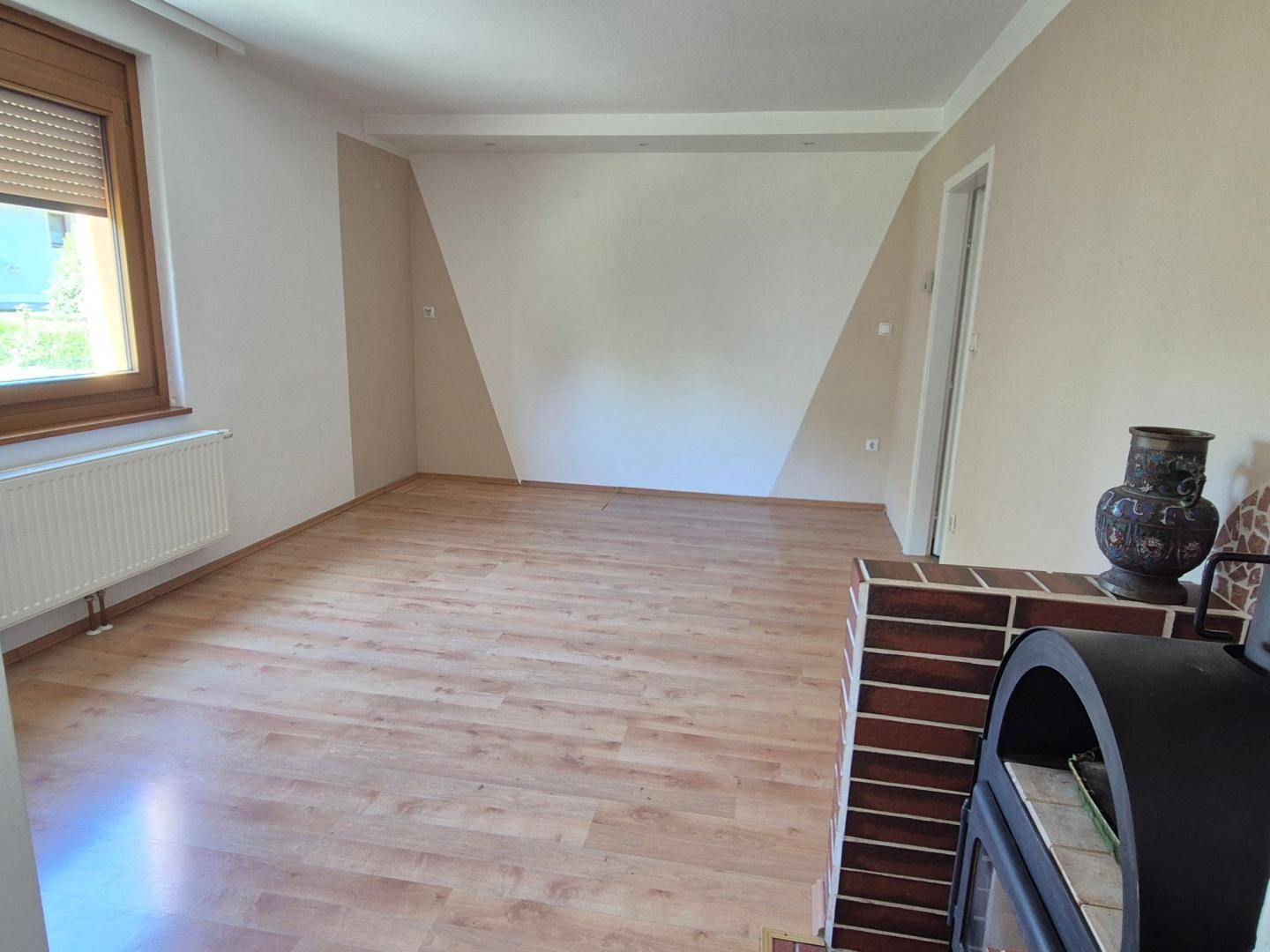 Helles Wohnzimmer mit Laminatboden, Heizkörper unter dem Fenster und einem Holzofen im Vordergrund.