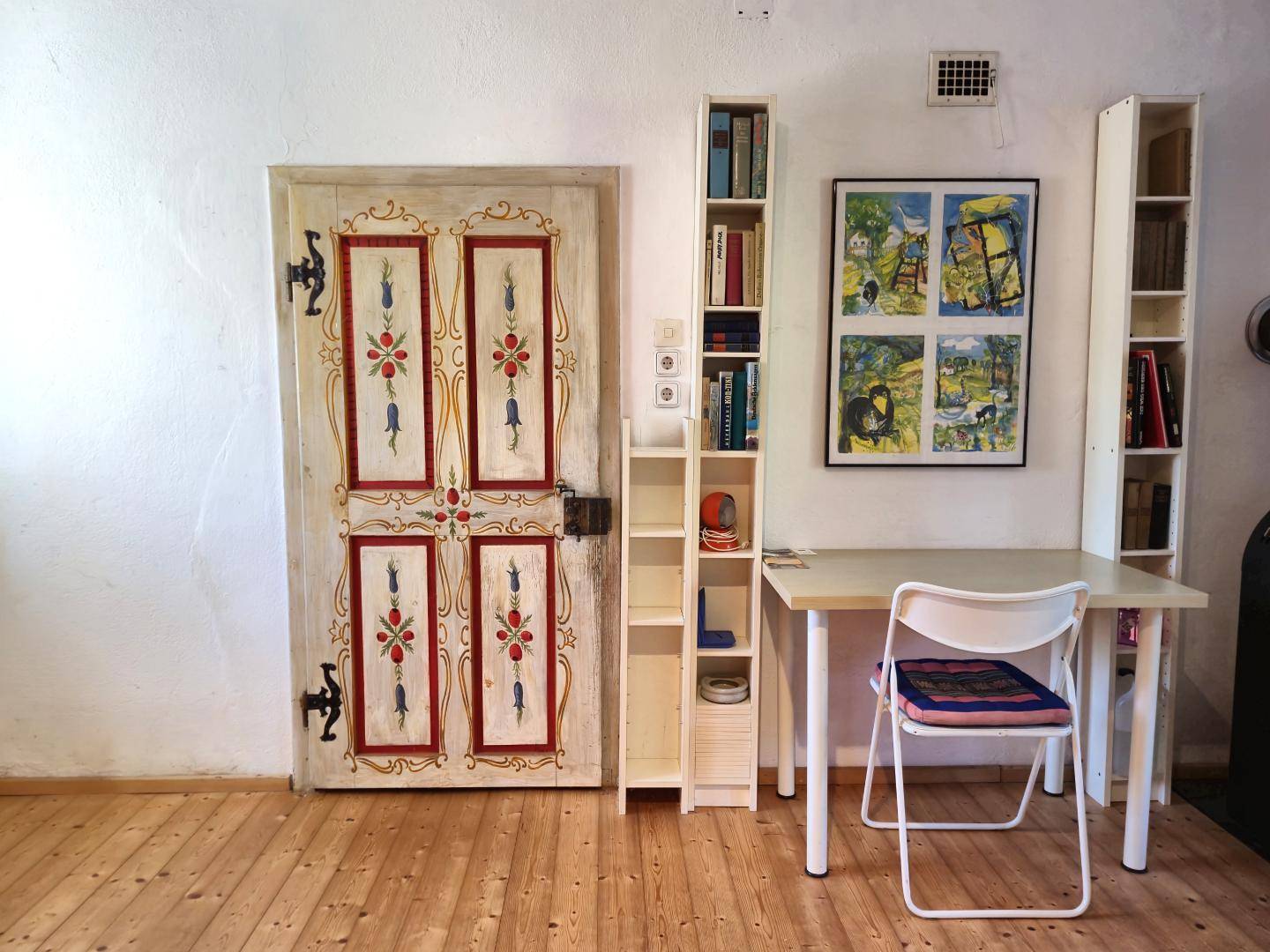 Arbeitsbereich mit Schreibtisch, Bücherregalen und einer kunstvoll bemalten Holztür auf Holzboden.