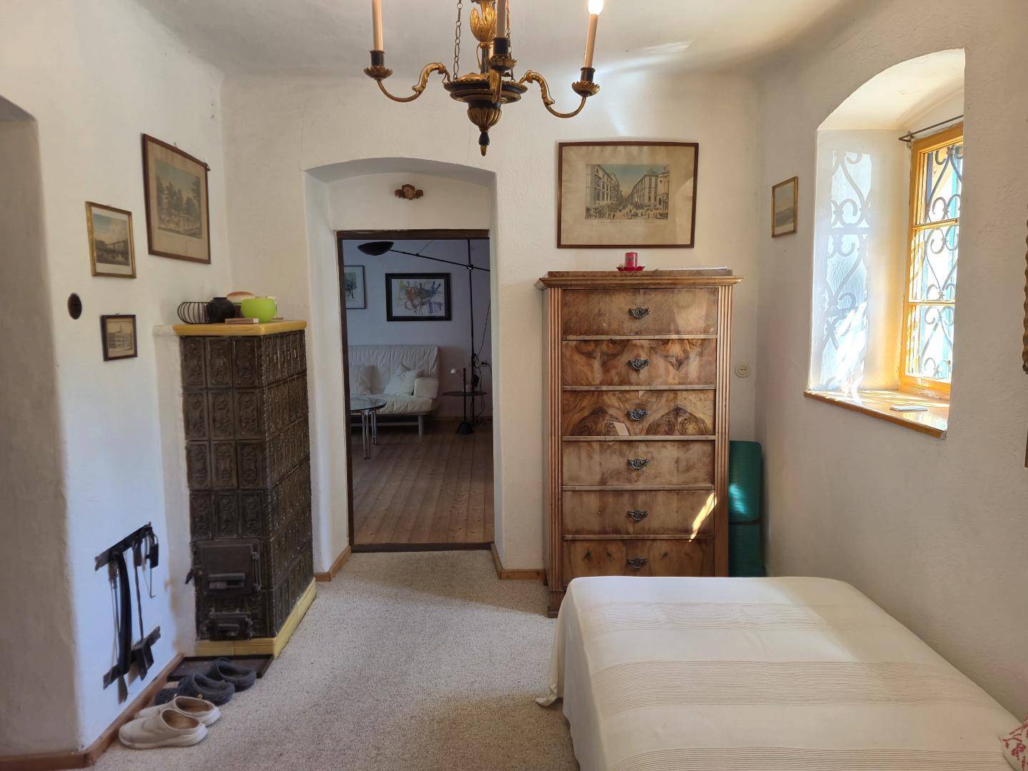 Helles Schlafzimmer mit traditionellem Kachelofen, antiker Kommode und Blick in den nächsten Raum.