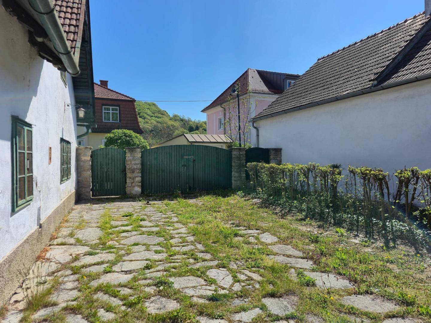 Schotterweg führt zu einem grünen Holztor, umgeben von Hecken und weiteren Gebäuden.