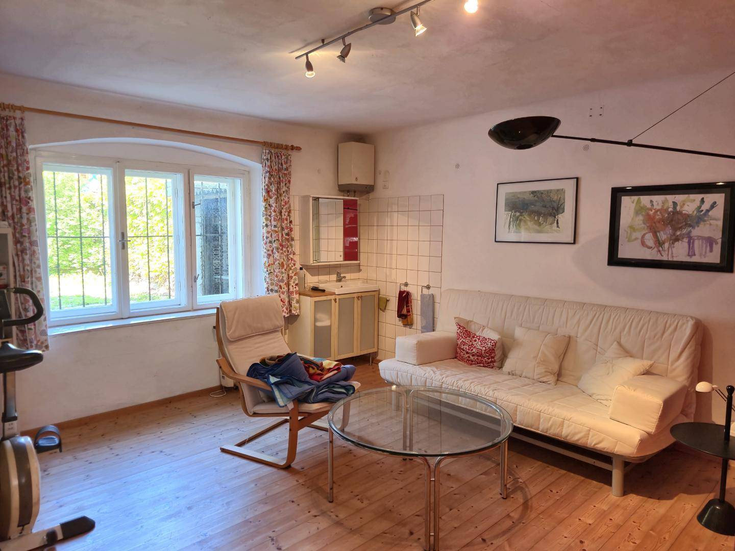 Wohnzimmer mit Sofa, Fitnessgeräten und einer kleinen Küchenzeile unter einem Fenster.