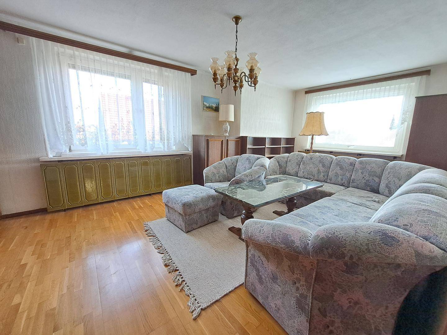 Gemütliches Wohnzimmer mit großem Ecksofa, Holzboden und zwei Fenstern, die den Raum hell erscheinen lassen.