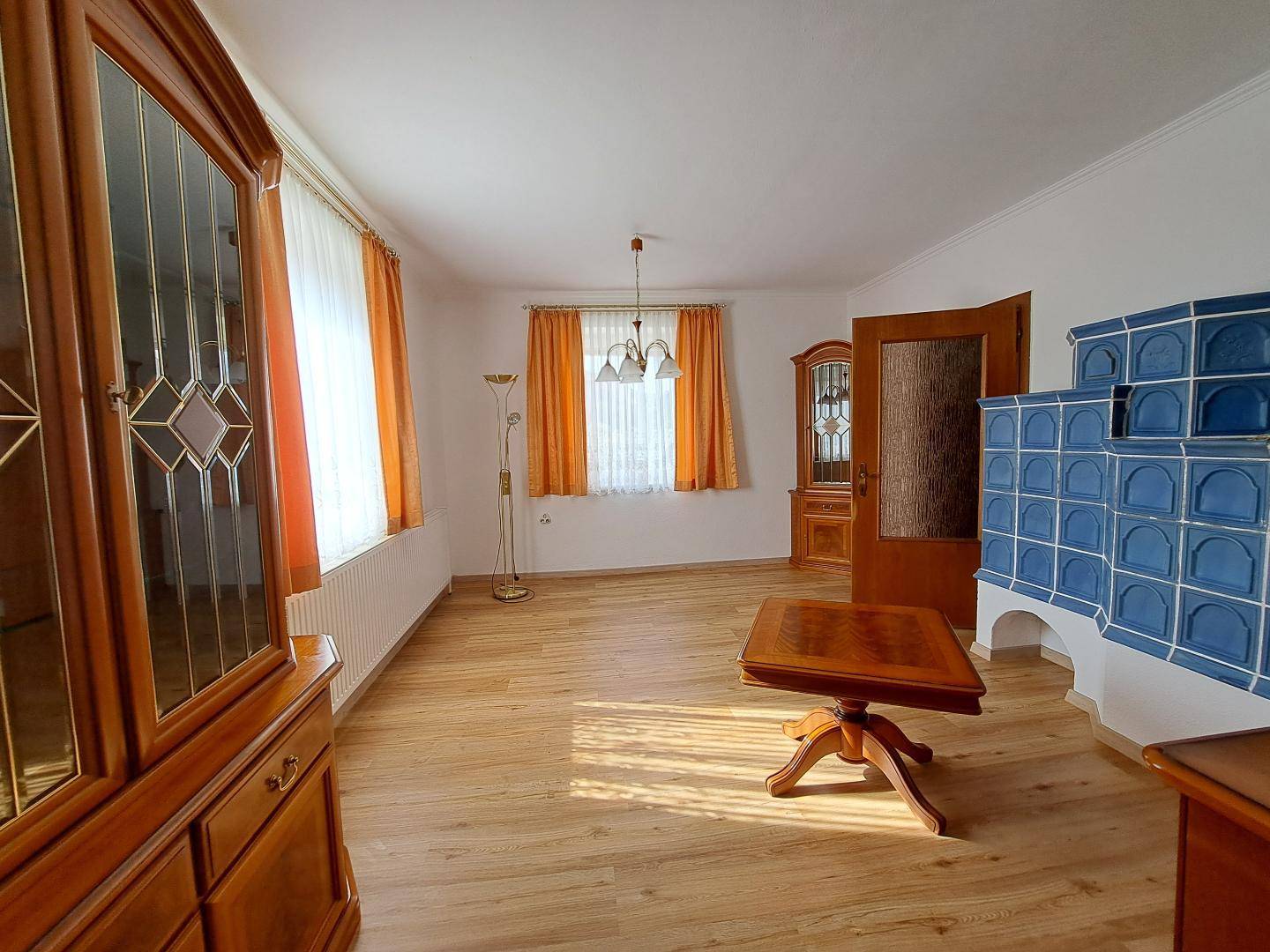 Helles Wohnzimmer mit Holzboden, einem traditionellen blauen Kachelofen und klassischen Holzmöbeln.