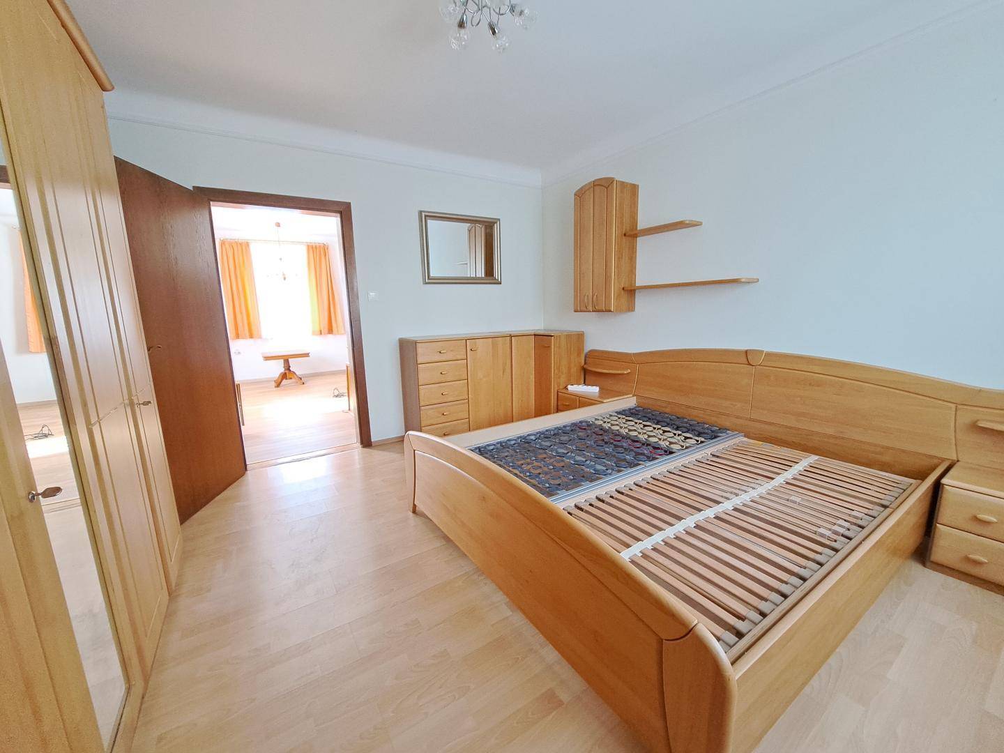 Schlafzimmer mit hellem Holzbett, passendem Kleiderschrank und Laminatboden, durch die Tür ist ein weiterer Raum sichtbar.