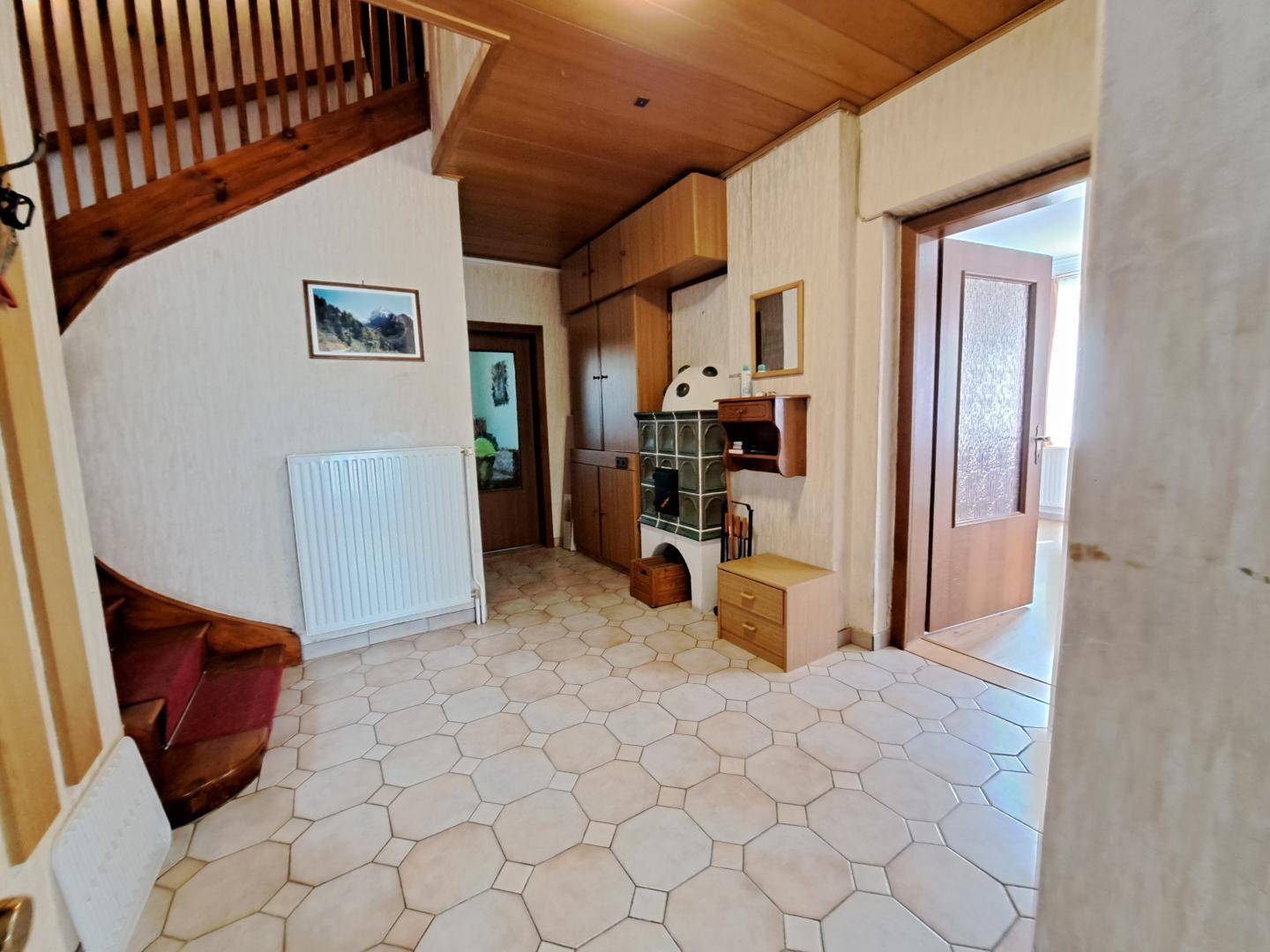 Geräumiger Flur mit gemustertem Fliesenboden, Holztreppe und einem kleinen Kachelofen als Blickfang.
