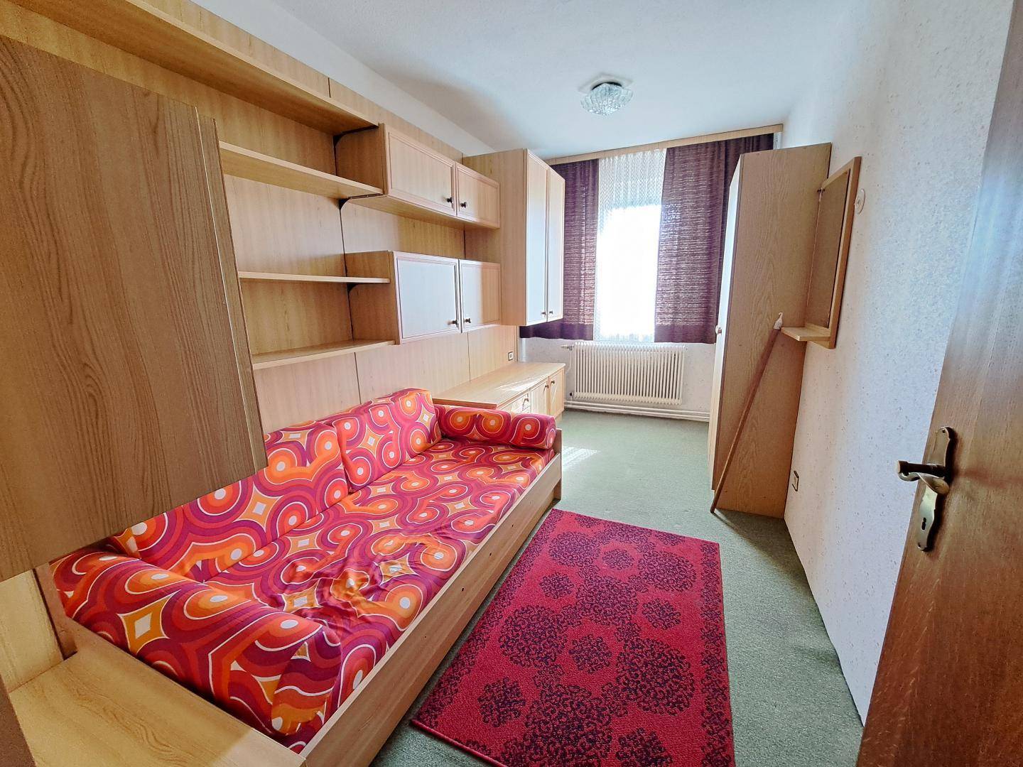Helles Zimmer mit Tagesbett, integrierten Holzregalen und einem farbenfrohen Teppich, ideal als Gästezimmer.