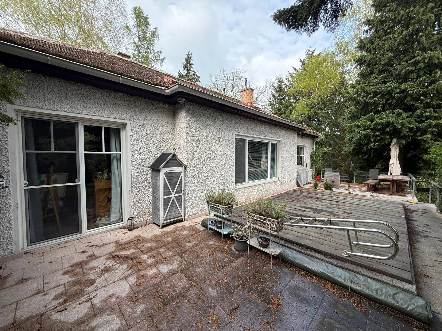 Gepflasterte Terrasse am Haus mit einer Abdeckung, umgeben von Bäumen und Grünflächen.