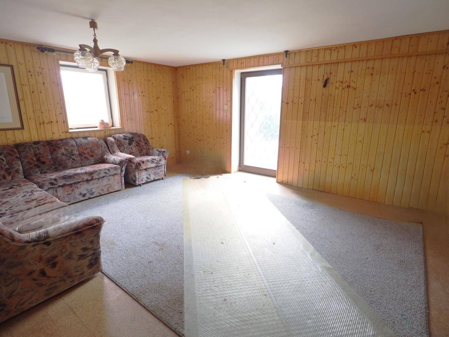 Rustikaler Wohnraum mit Holzvertäfelung, Teppichboden und Zugang zum Außenbereich.