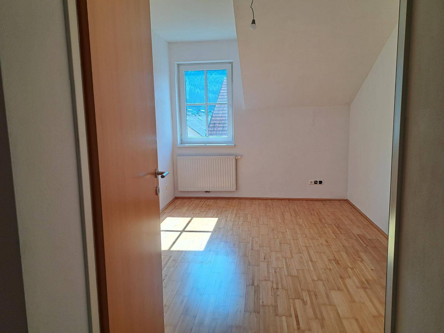 Gemütliches Zimmer mit Holzboden und Dachschräge, gut beleuchtet durch ein Fenster.