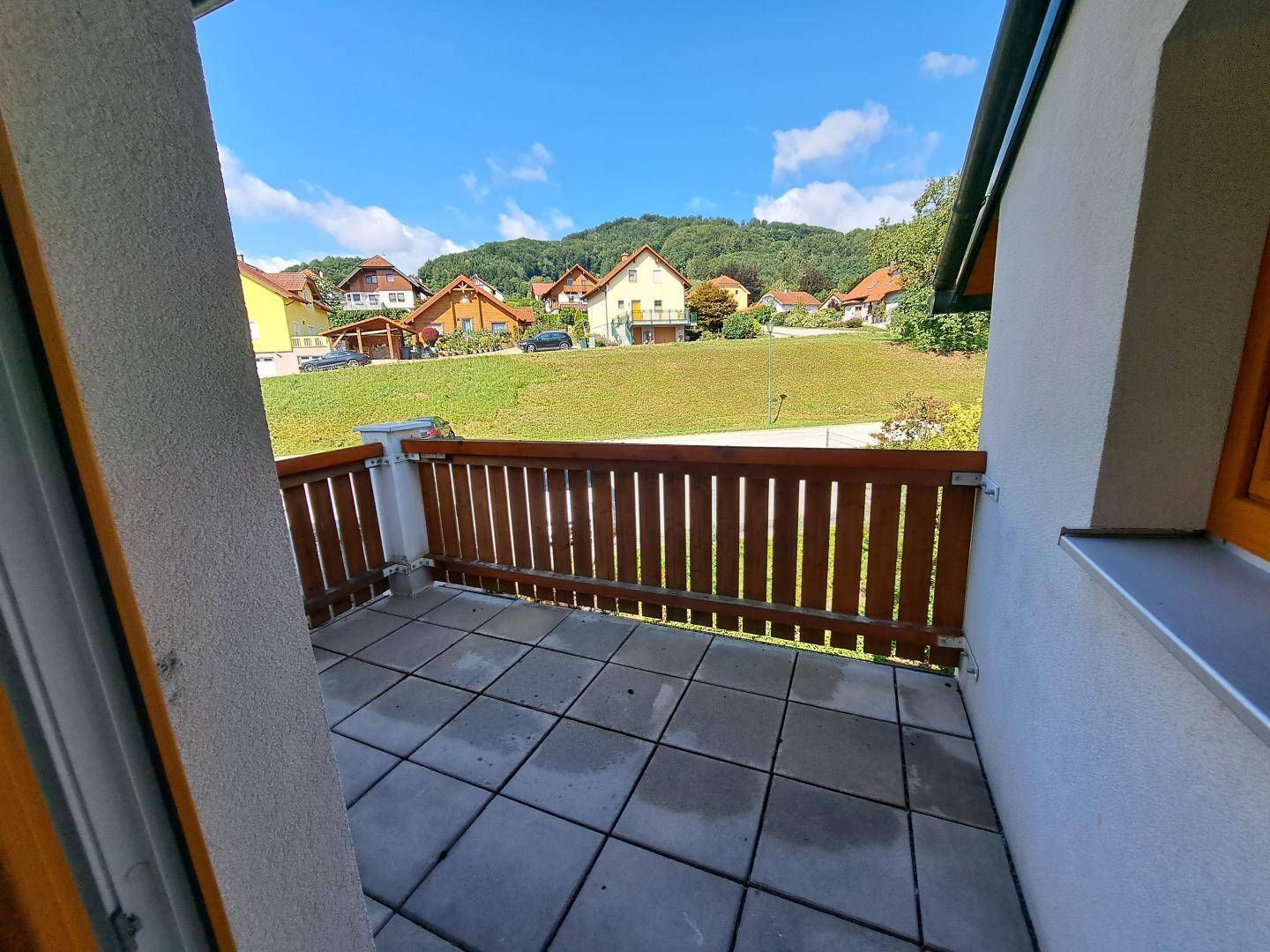 Sonniger Balkon mit Holzzaun und Blick auf die grüne Landschaft und umliegende Häuser.