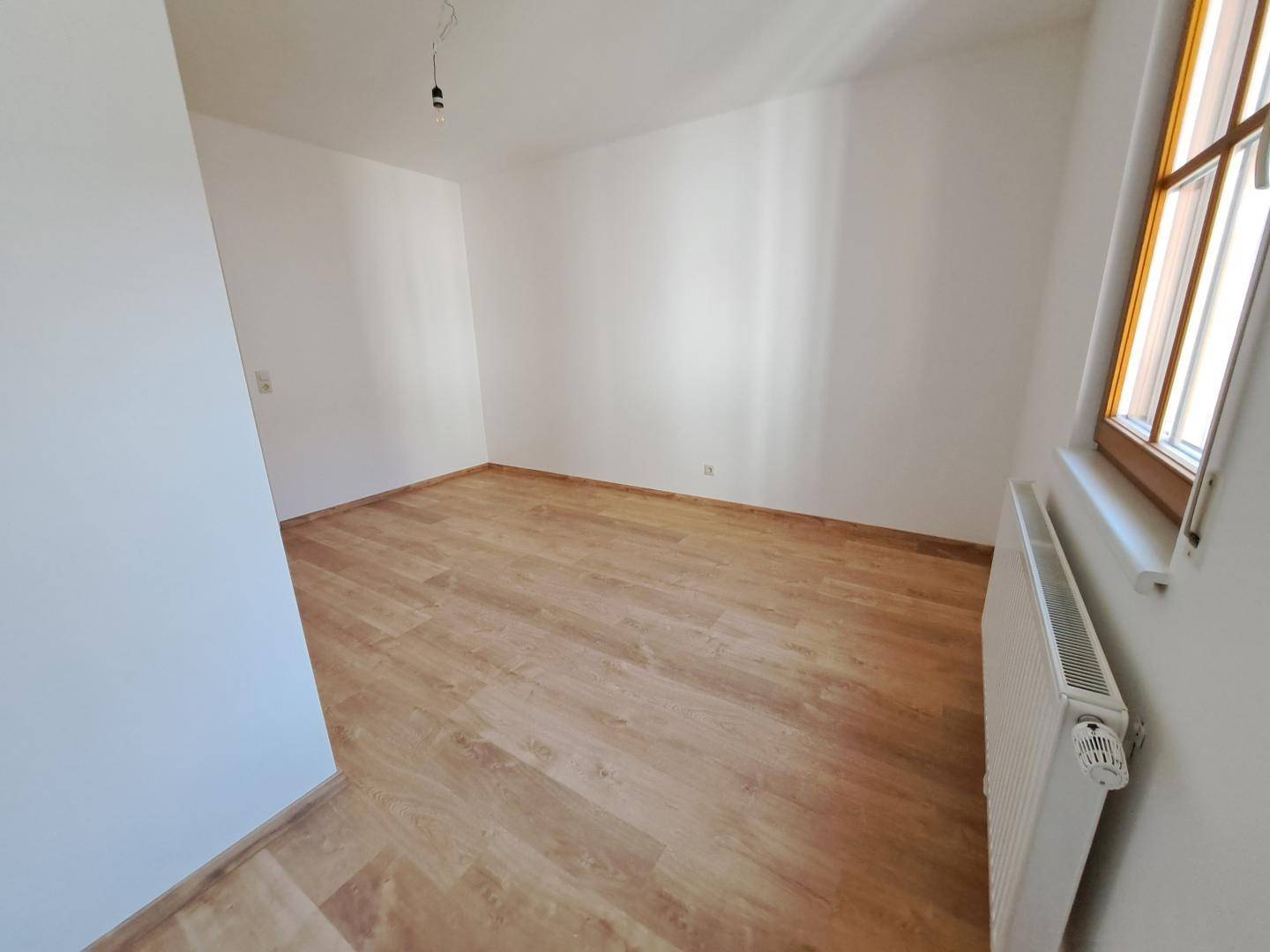 Helles Zimmer mit Laminatboden und Heizkörper unter dem Fenster.