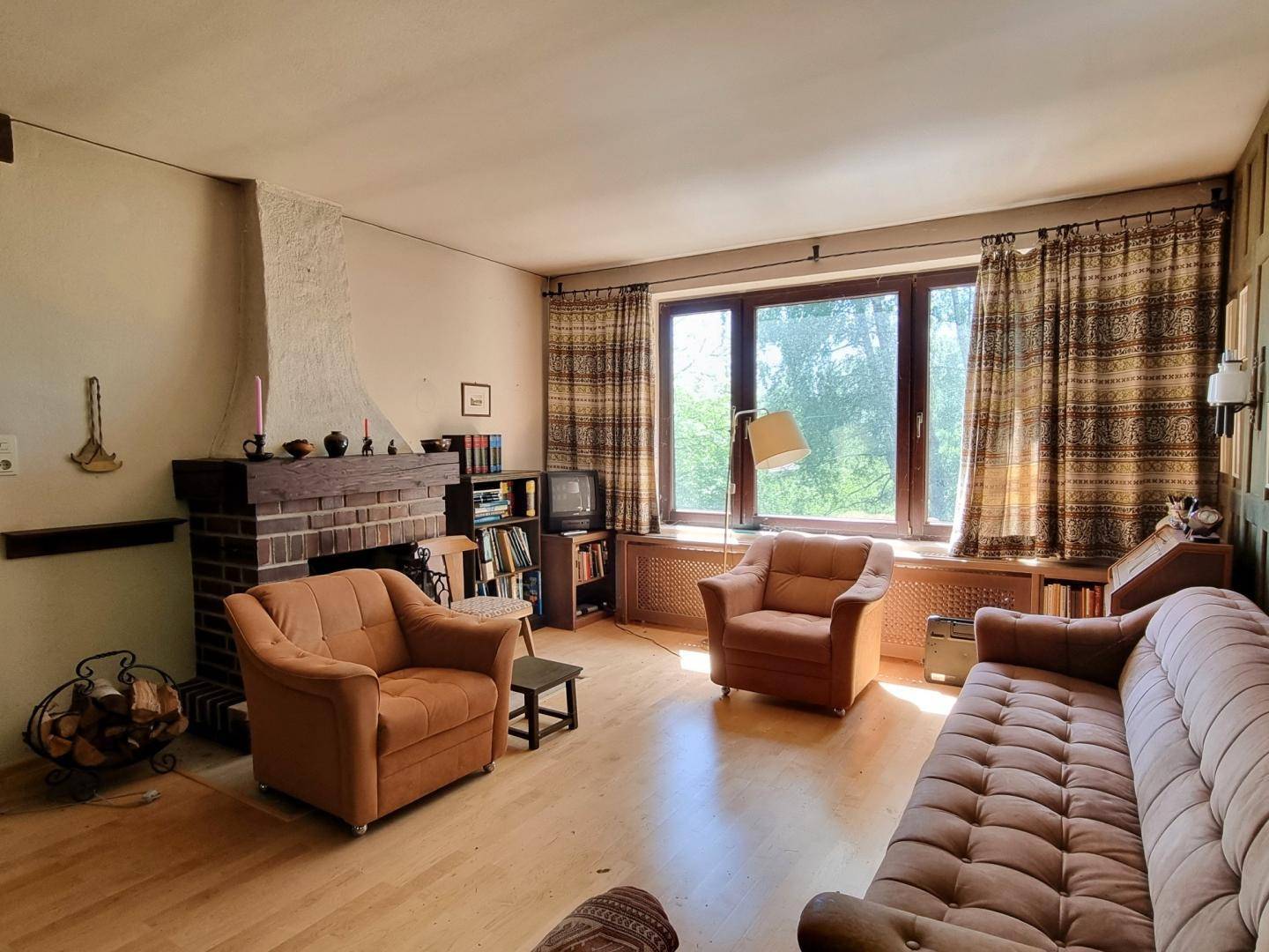 Wohnzimmer mit traditionellem Ziegelkamin, Holzboden und großen Fenstern mit Blick ins Grüne.