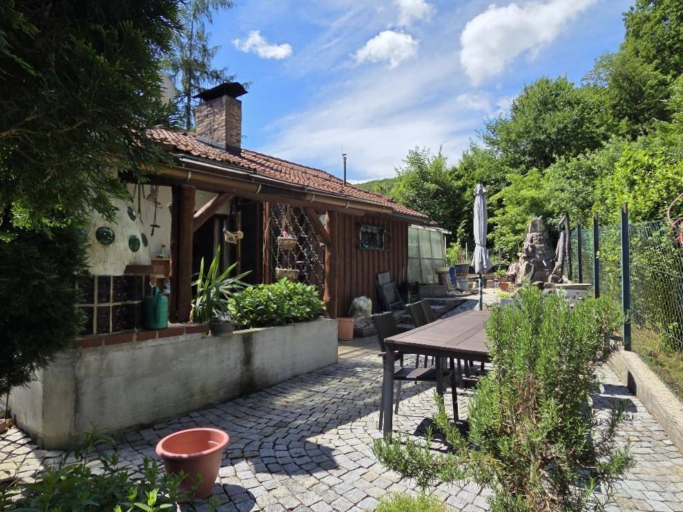Gemütliche gepflasterte Terrasse mit Esstisch und Stühlen, umgeben von üppigem Grün und Bäumen.