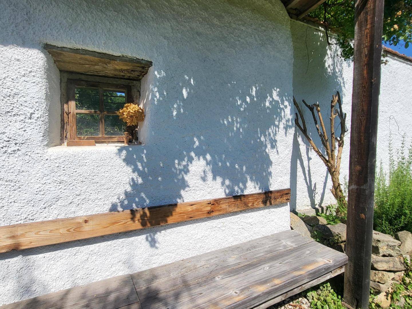 Detailansicht einer weißen, strukturierten Wand mit kleinem Holzfenster und rustikaler Holzbank.