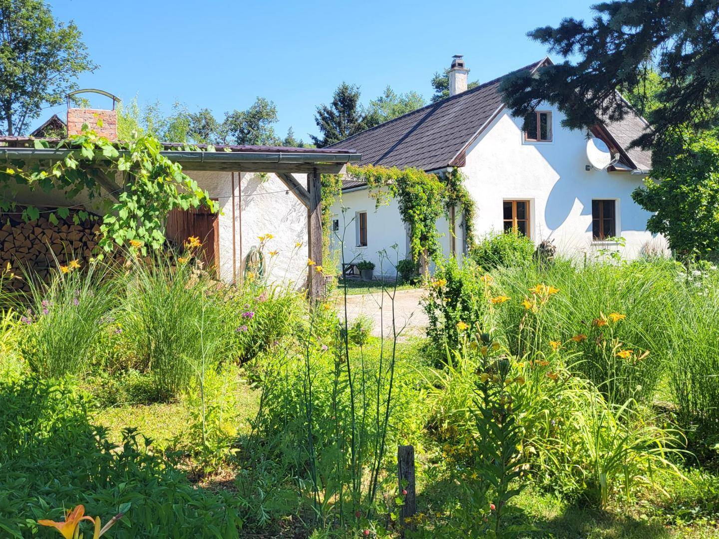 Weißes Haus mit braunem Dach, umgeben von einem üppigen Garten mit Blumen und Pflanzen.